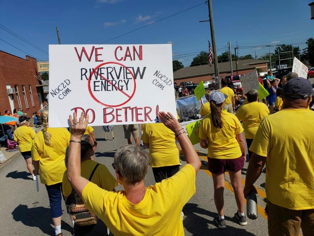 Protestors in yellow shirts holding a sign saying "We can do better" with "Riverview EnergyEnergy" having a red circle and slash around it and "NOC2D.com" written on the sides